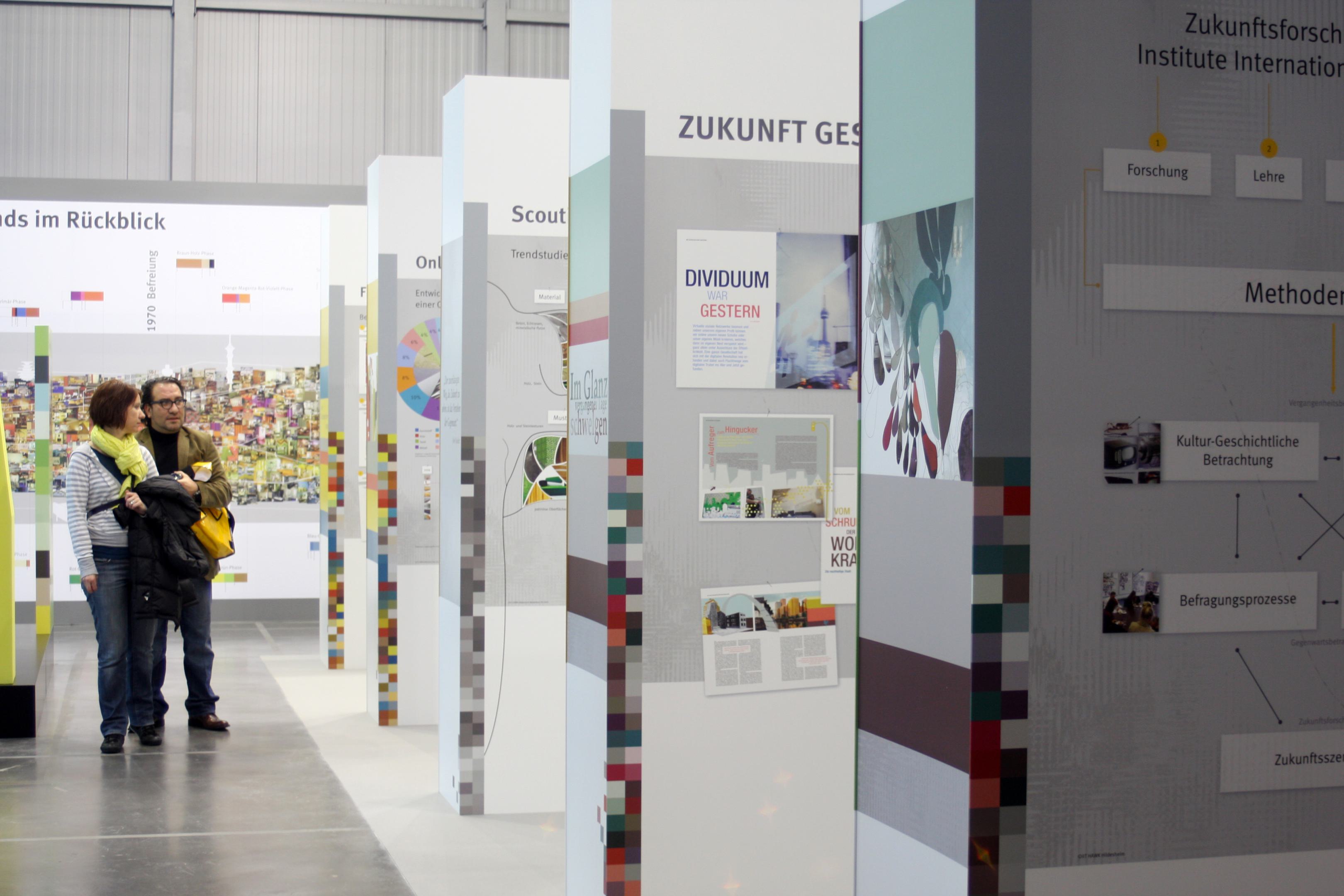Messestand mit farbigen Stelen und einer Wand mit den Farb- und Materialzyklen in einer kleinen Bildersammlung f&uuml;r den Innenraum