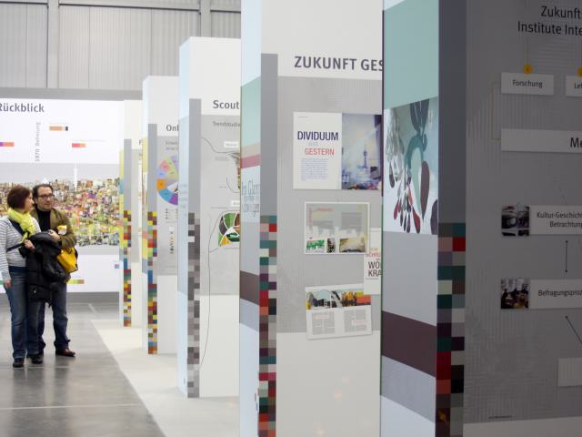 Messestand mit farbigen Stelen und einer Wand mit den Farb- und Materialzyklen in einer kleinen Bildersammlung f&uuml;r den Innenraum